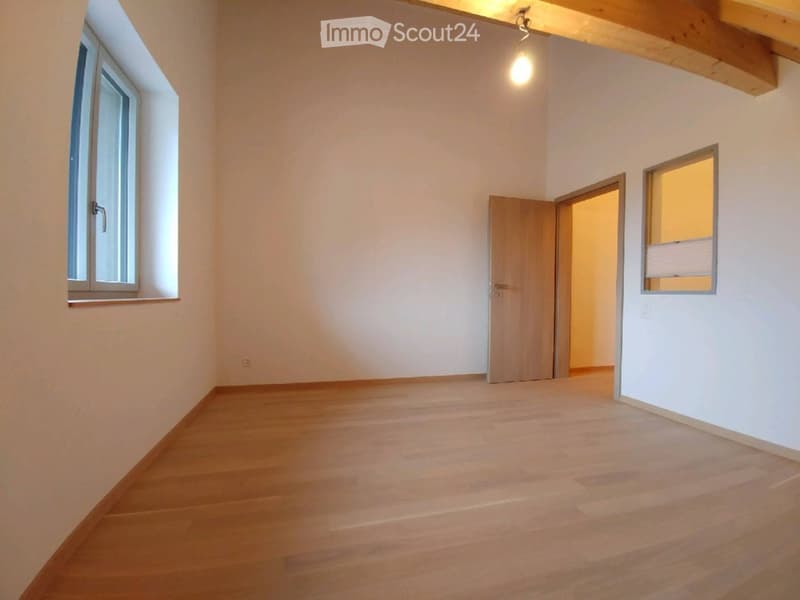 Empty room with wooden flooring, white walls, and exposed wooden beams on the ceiling. Features two windows and an open doorway leading to another space.