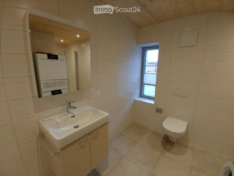 Modern bathroom with beige tiles, a white sink with a modern faucet, a large mirror, a toilet, and a small window providing natural light.