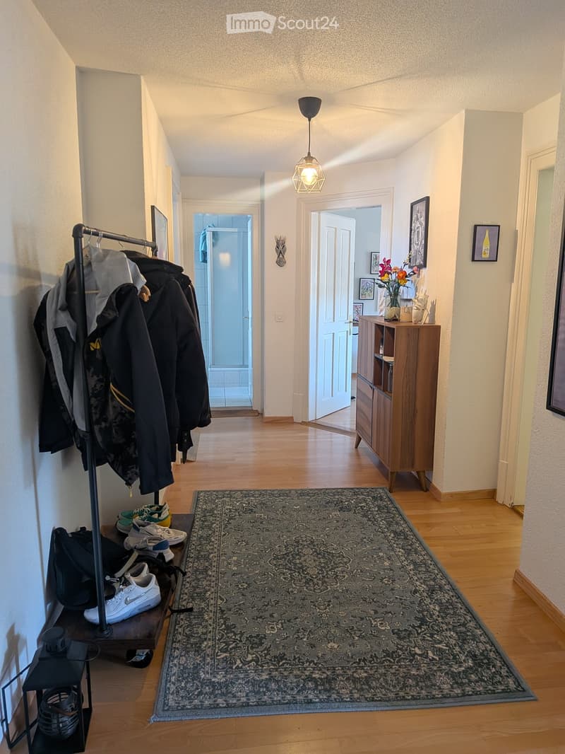 A well-lit hallway with wooden flooring, a patterned rug, coat rack with jackets, shoes, and a small wooden cabinet with decorative items and a vase of flowers.
