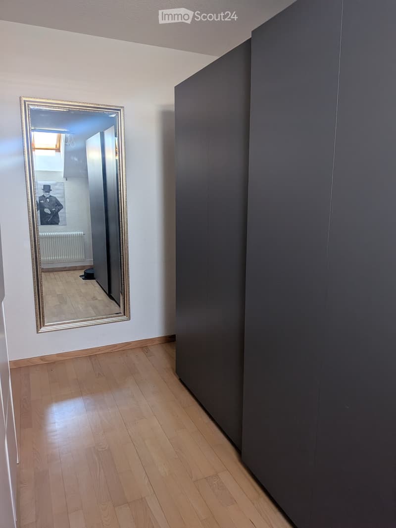 Empty hallway with wooden flooring, white walls, and a large mirror on one side. The mirror reflects part of an adjacent room with a radiator and a framed picture.