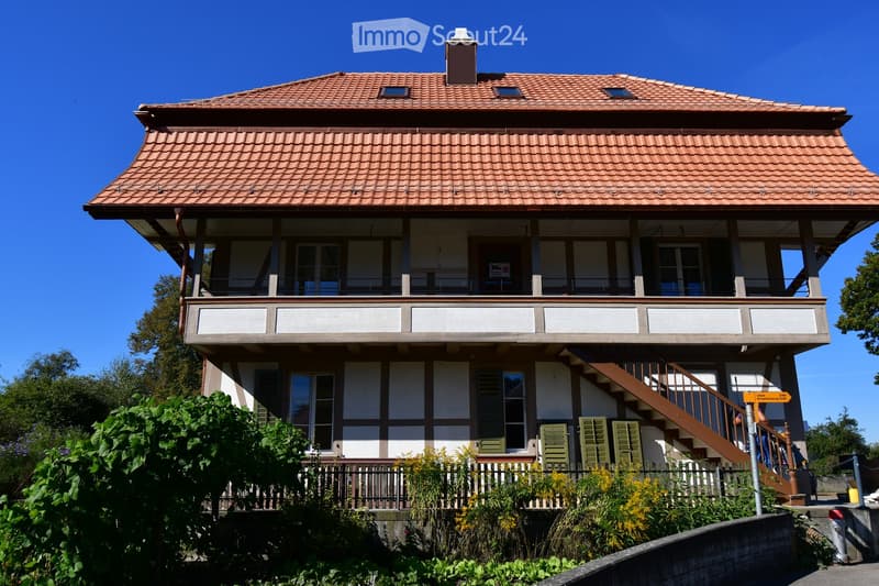 Two-story house with a red-tiled roof, white and grey exterior, multiple windows, and a balcony on the upper floor.