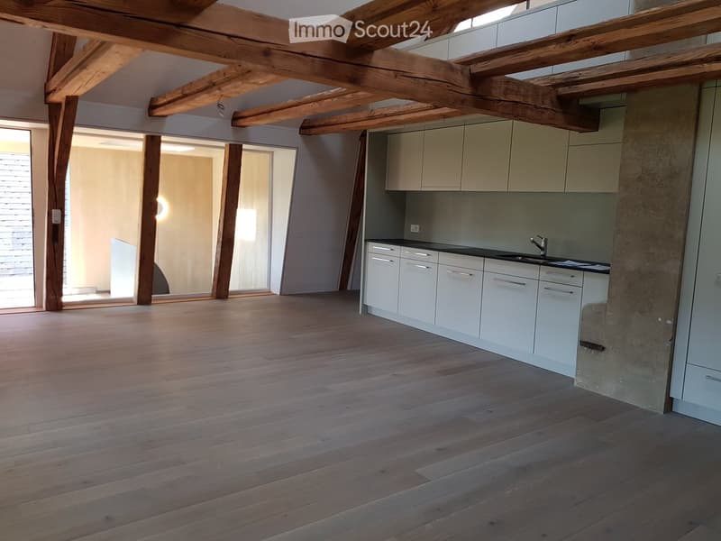 Open-plan space with exposed wooden beams and a modern kitchen area featuring white cabinets and a black countertop. The floor is made of light wooden planks, and there is a large window providing natural light.