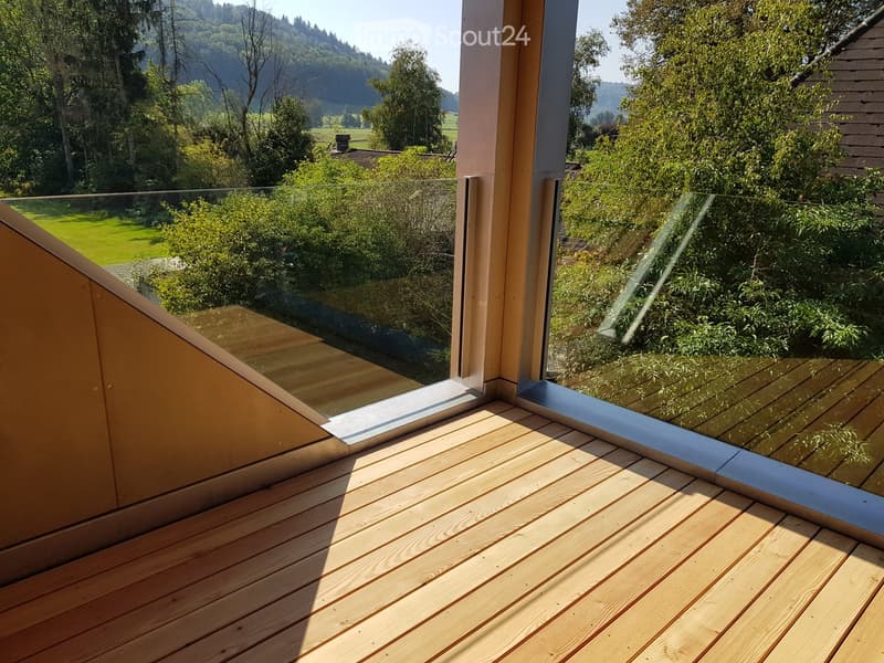 Wooden deck with glass railing, overlooking a green landscape with trees and hills.