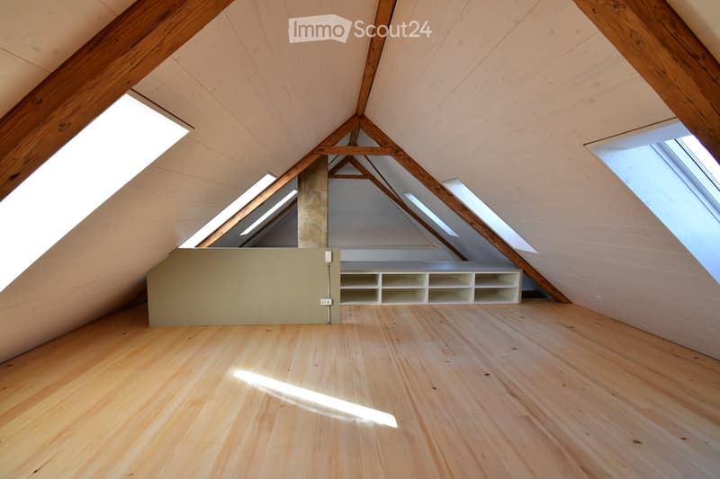 An empty attic space with wooden floors, exposed wooden beams, and skylights allowing natural light to enter. The room features built-in white shelving units along one wall.