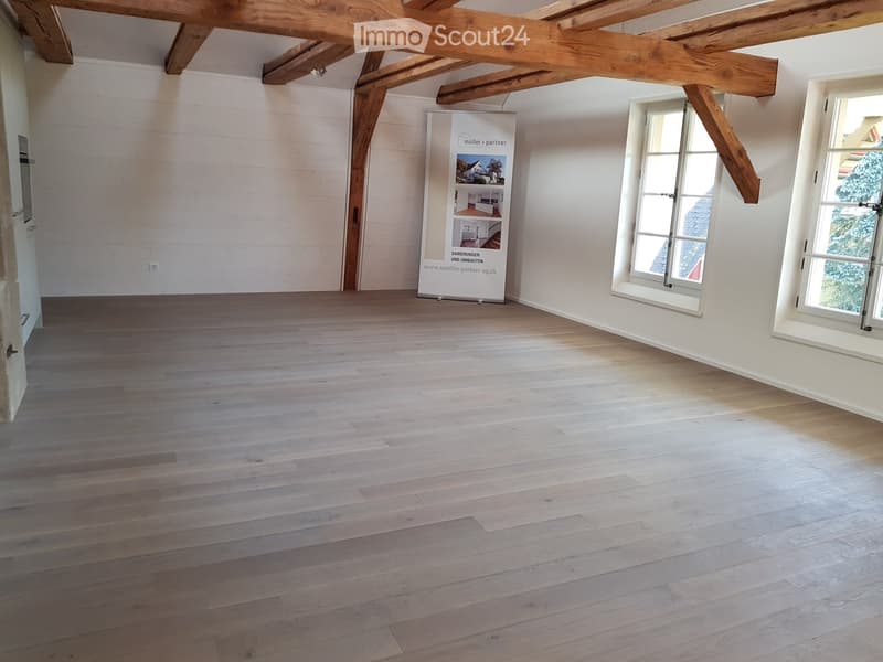 Empty room with wooden floor, white walls, exposed wooden beams, and two windows with white frames.