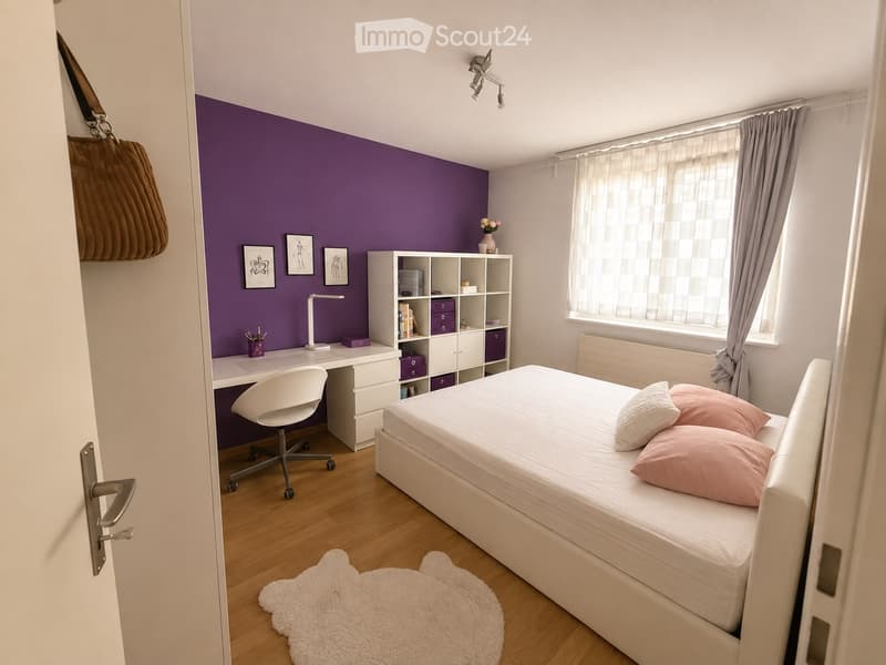 Modern bedroom with a white bed, pink pillows, a white desk with a chair, a white shelving unit, and a purple accent wall.