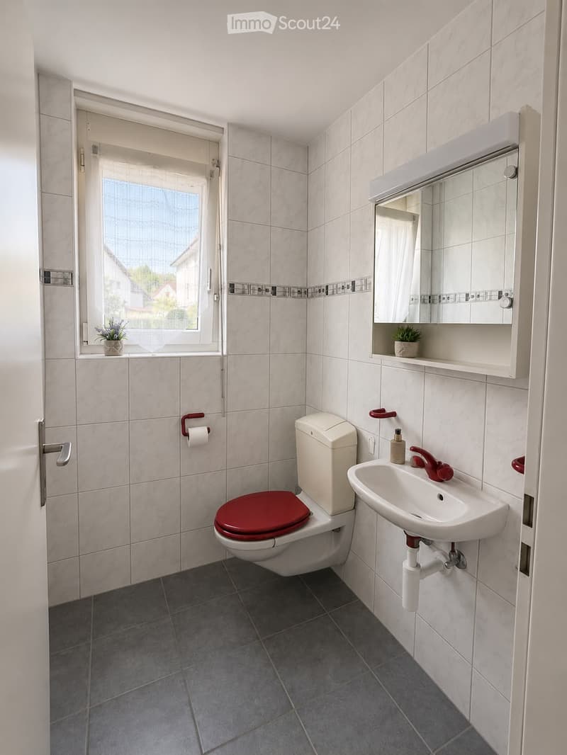 Modern bathroom with white tiles, a red toilet seat, a white sink with red faucet, a mirrored cabinet, and a small window with a plant on the sill.