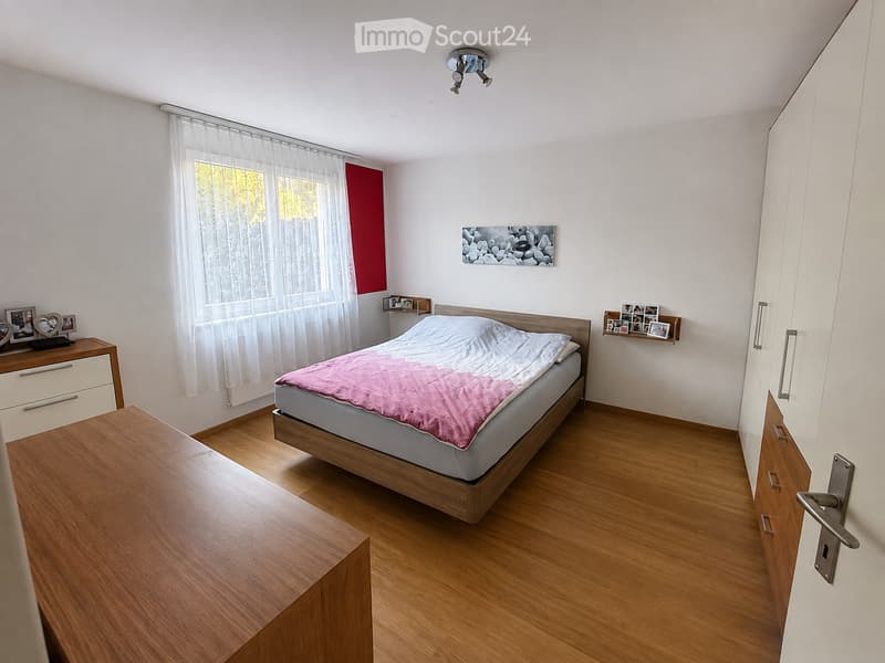 Bedroom with wooden floor, large bed with pink blanket, nightstand, mirror, picture frames, and built-in cabinet.