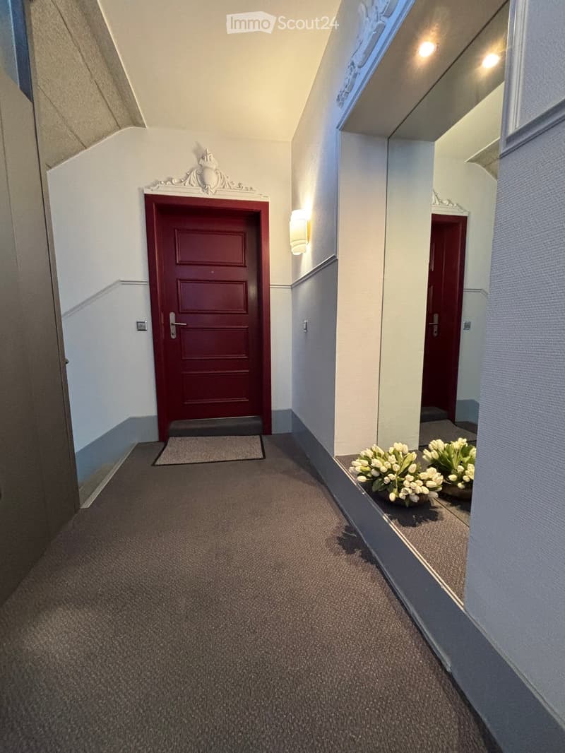 Carpeted hallway with two red doors, wall-mounted light fixture, and floral arrangements on the right side.