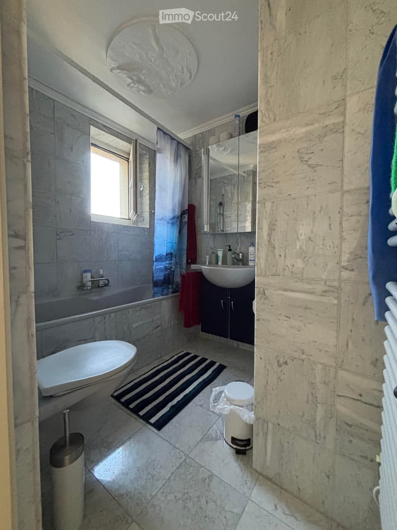 Modern bathroom with marble tiles, a bathtub, a sink, a mirror, a cabinet, and a towel hanging on the wall. The room features a striped rug and a small window letting in natural light.