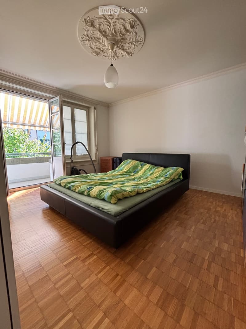 Modern bedroom with wooden flooring, large window with blinds, black leather bed frame, green and yellow bedding, ornate ceiling light fixture, and a small nightstand.