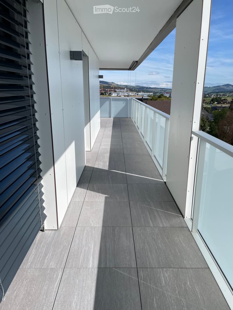 Modern balcony with grey tiled floor, white walls, glass railing, and scenic view of the landscape.