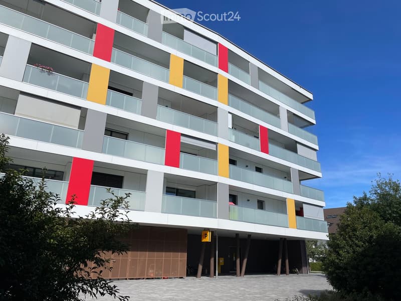 Modern multi-story apartment building with balconies, colorful panels, and a parking area.