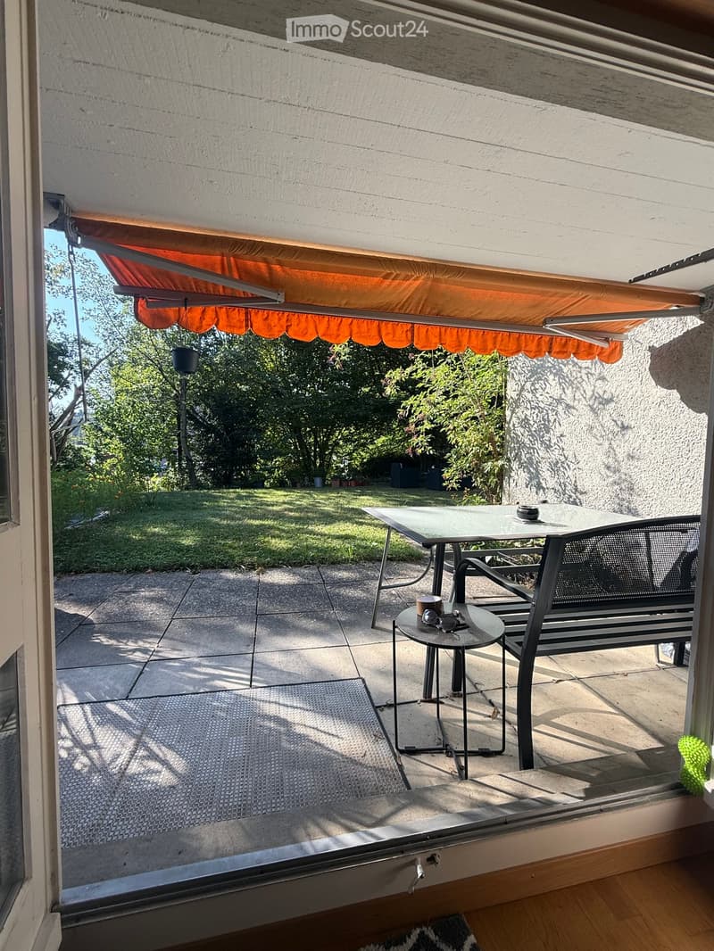 Balcony with a glass table, two chairs, and a small round stool, featuring an orange awning and a view of a green garden.