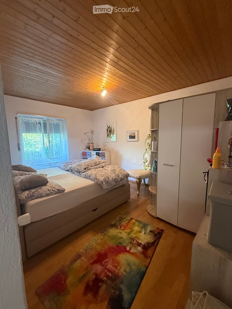 Cozy bedroom with wooden slanted ceiling, large window with sheer curtains, double bed with patterned bedding, colorful rug, wardrobe, small table with decorative items, and a chair near the window.