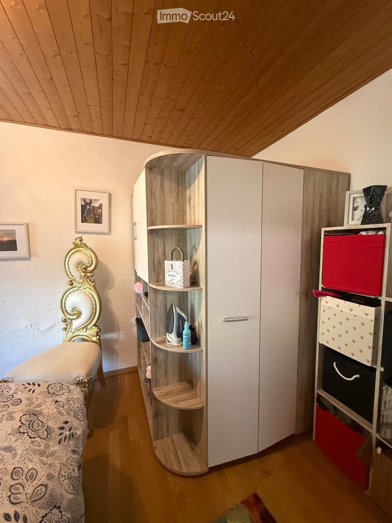 Cozy bedroom with a wooden slanted ceiling, a white bed with ornate headboard, a large wardrobe with shelves, and a small shelf holding an iron and other items.