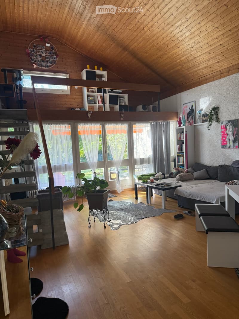 A spacious, well-lit living room with wooden floors, a raised ceiling, and large windows. Features include a sofa, coffee table, stools, a plant, shelves, and artwork.