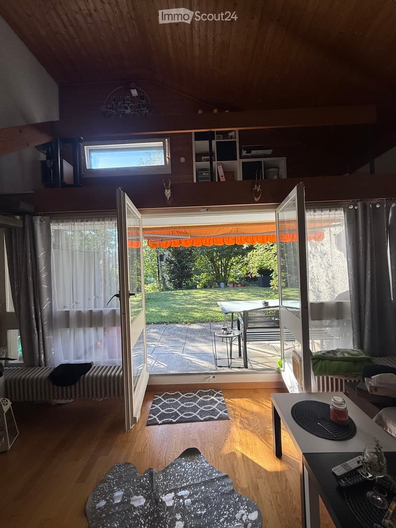 Bright living space with wooden floors, large windows, and an open sliding door leading to a patio with outdoor furniture.