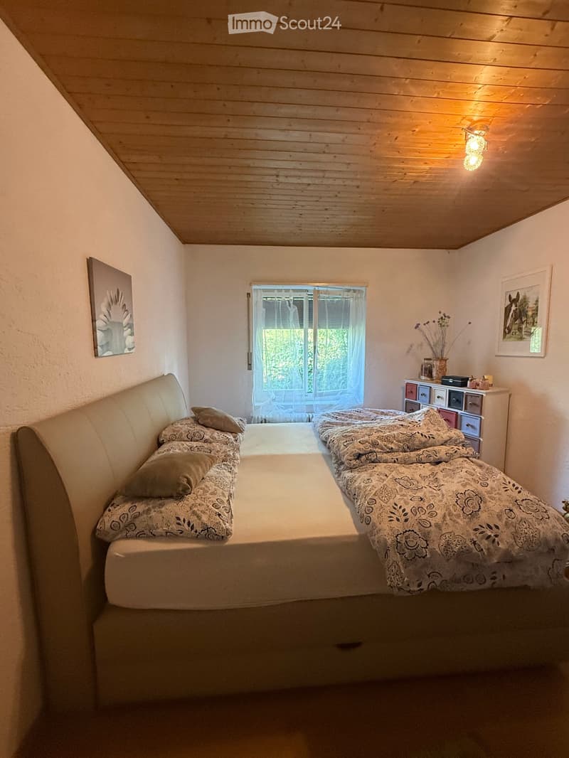 Cozy bedroom with a large bed, a small sofa, nightstand, and artwork on the walls. The room features wooden ceilings and a window with sheer curtains.