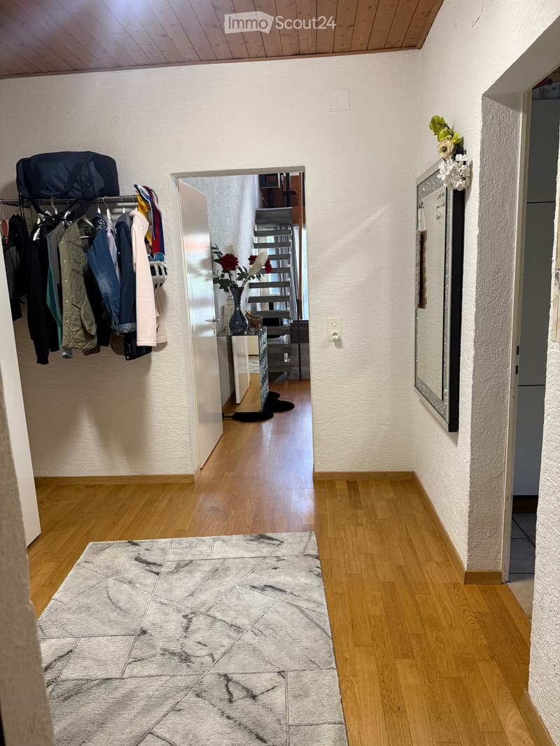 Hallway with wooden flooring, a rug, a coat rack with jackets, a small table with a vase of flowers, and a mirror on the wall.