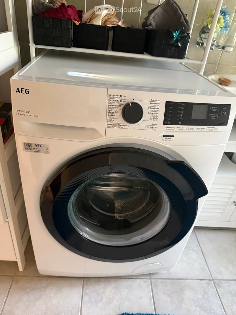 A modern white washing machine with a digital control panel, placed on a tiled floor with a shelf above holding folded clothes and decorative items.