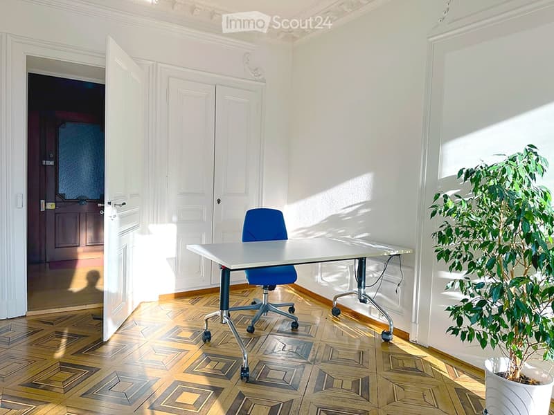 Bright room with wooden parquet flooring, white walls, a white table with a blue chair, and a potted plant near the window.