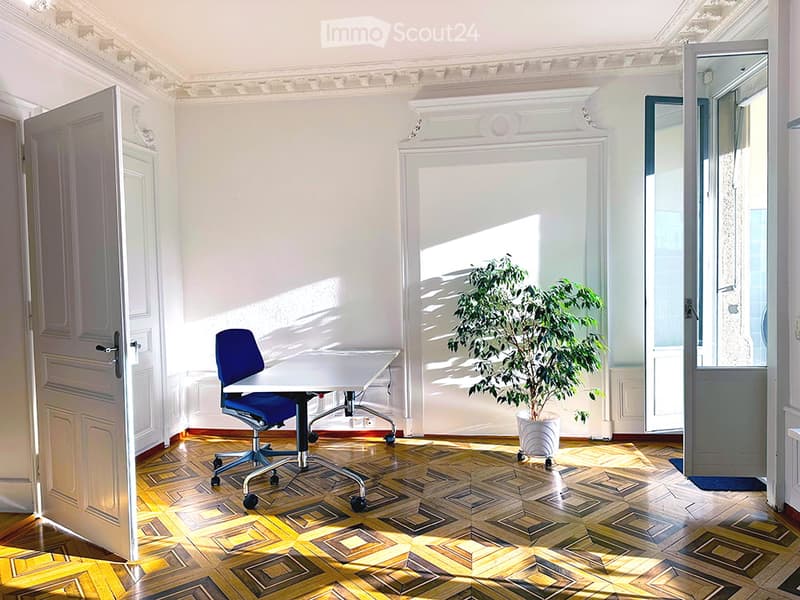 Bright room with wooden parquet flooring, white walls, and large windows. Features a blue office chair, white desk, and a potted plant near the window.