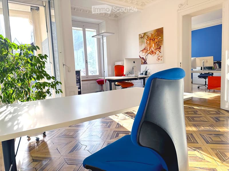 Modern office space with wooden parquet flooring, large windows, a white table, blue chair, laptop, and a plant in the corner.