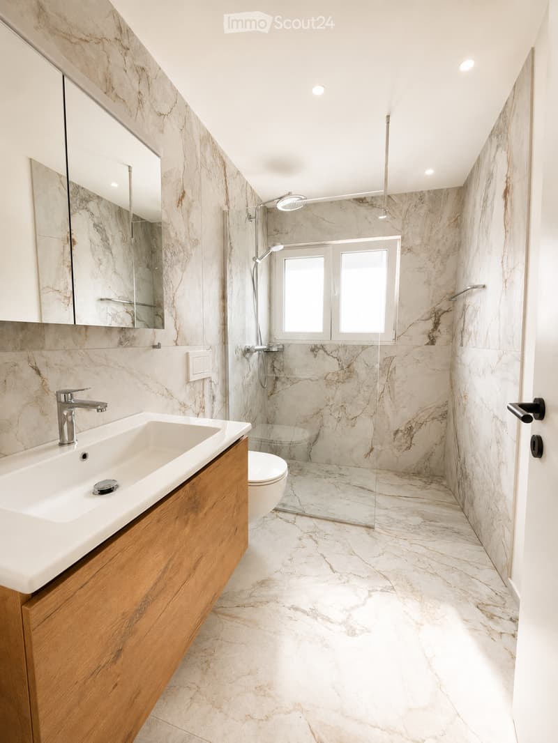 Modern bathroom with marble walls and floor, a large sink with wooden cabinet, a toilet, and a glass-enclosed shower area with a modern showerhead.