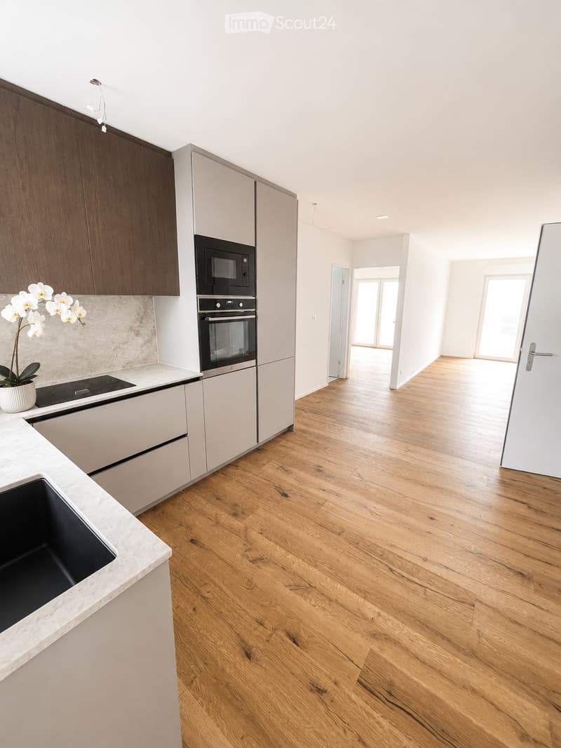 Modern kitchen with light wooden flooring, white and grey cabinets, a built-in microwave and oven, a marble countertop, and a decorative flower pot on the counter.