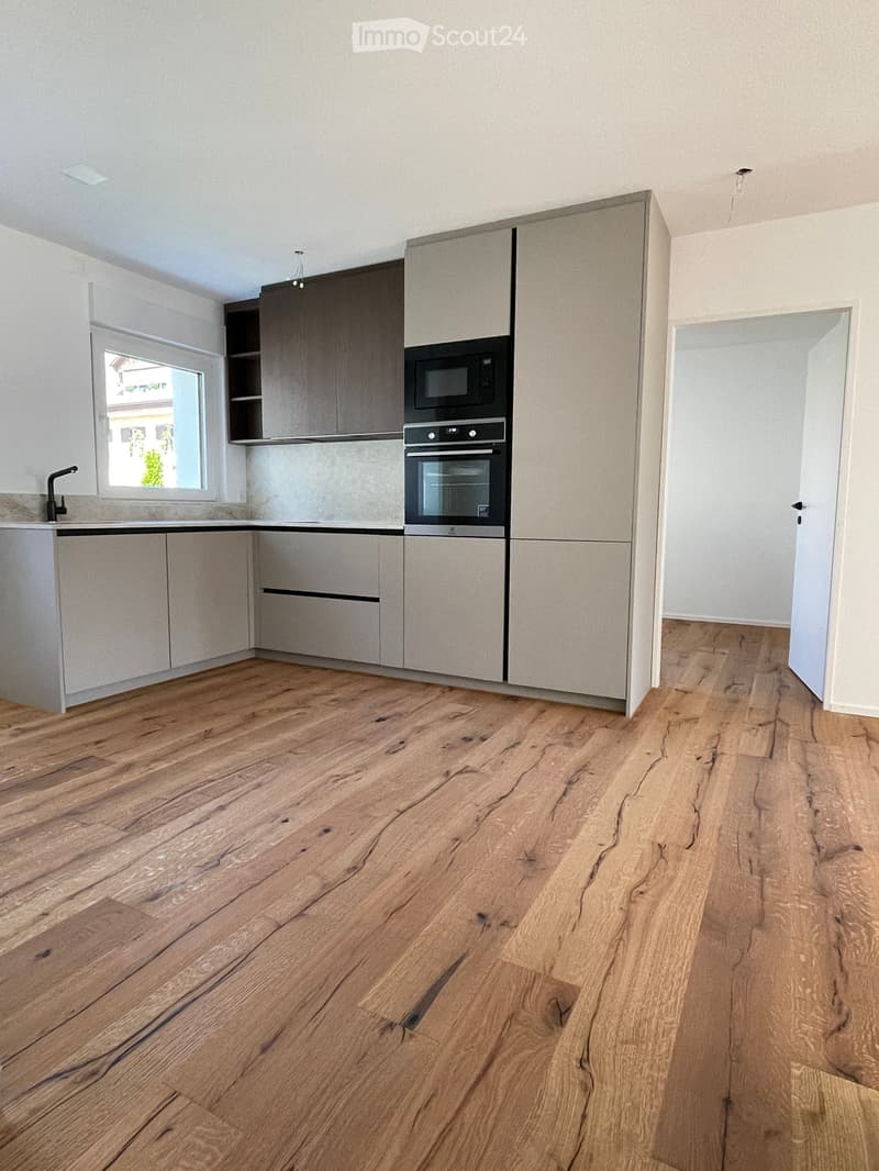 Modern kitchen with light grey cabinets, a microwave, an oven, and a faucet, featuring a wooden floor and a window with a view of greenery.