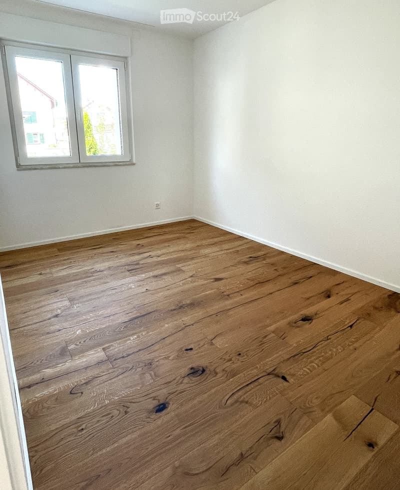Empty room with wooden flooring and white walls, featuring a large window with white frame and a visible electrical outlet on the wall.
