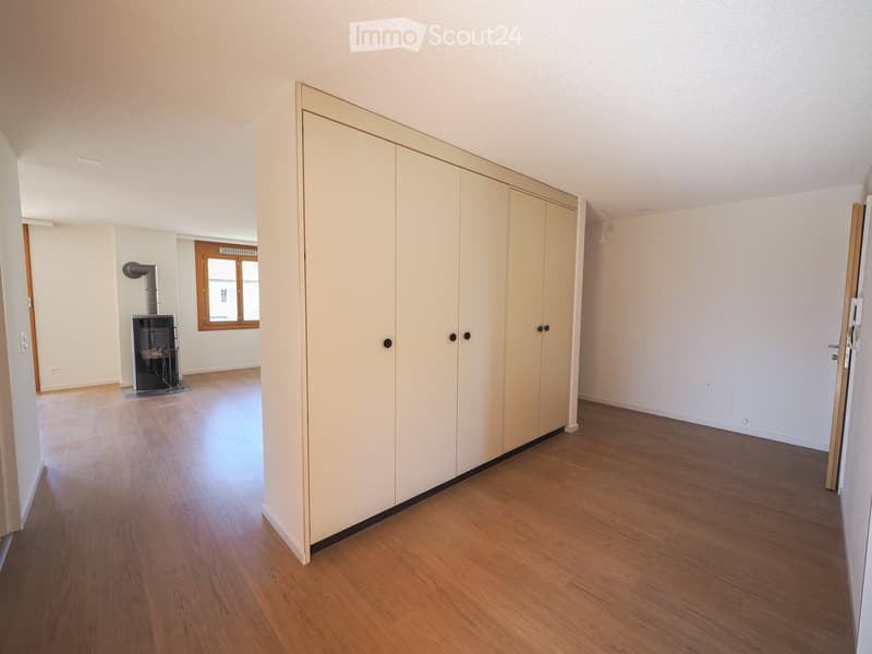 Empty room with wooden flooring, white walls, and a large built-in wardrobe. A fireplace is visible in the adjacent room through an open doorway.