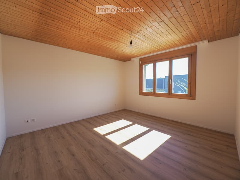 Empty room with wooden flooring, white walls, and a wooden ceiling. A large window with wooden frame allows natural light to enter.