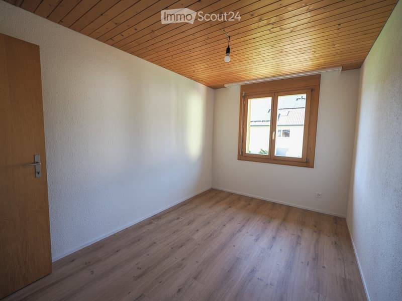 Empty room with wooden flooring, white walls, a wooden door, and a window with wooden frame. The ceiling is wooden with a single light fixture.