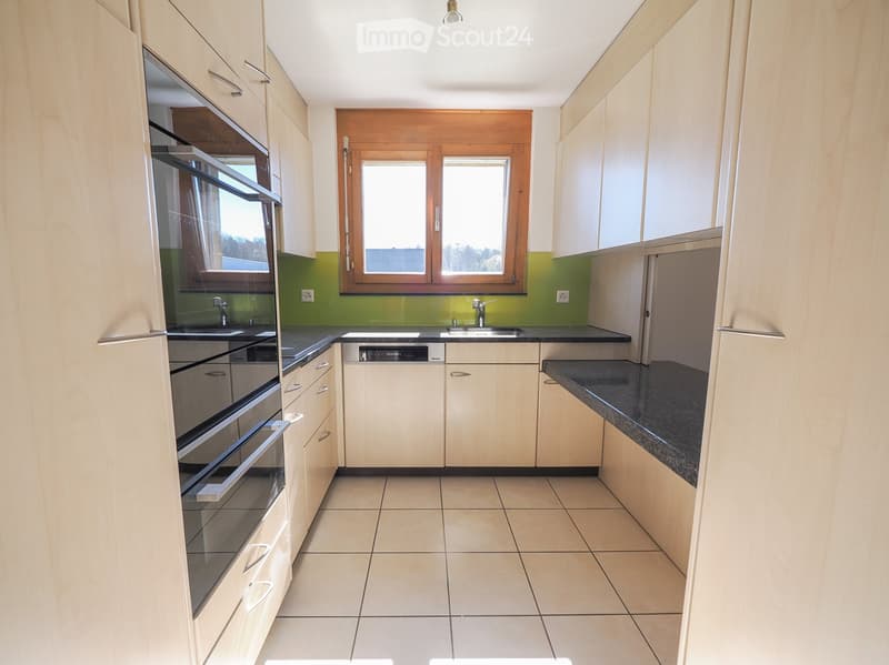 Modern kitchen with light wood cabinets, tiled floor, and stainless steel appliances including an oven and dishwasher. Green accent wall and large windows provide natural light.