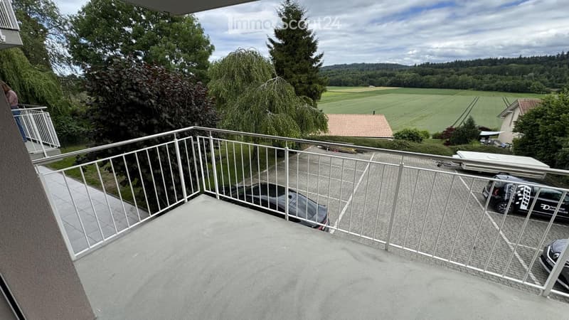 Balcony with metal railing overlooking a parking area and green landscape with trees and fields.