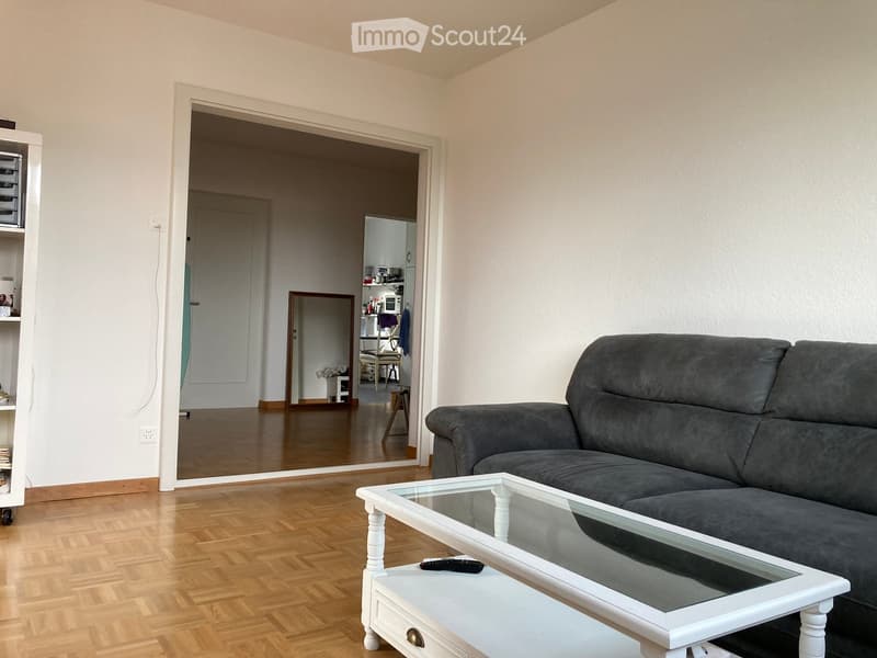 Living room with a grey sofa, a white coffee table, a mirror, and a shelf. The room has wooden flooring and white walls.