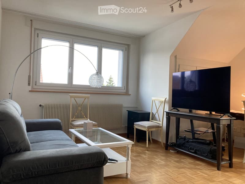 Living room with grey sofa, glass coffee table, white wooden chair, small stool, TV on a stand, and a floor lamp near the window.