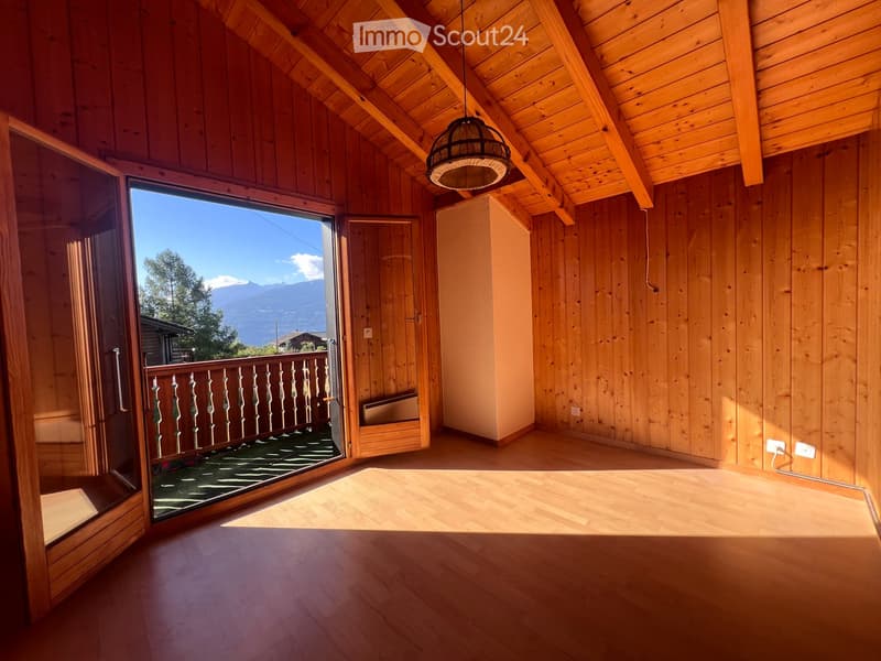 Bright, empty room with wooden walls and ceiling, large window offering a scenic mountain view, and a balcony accessible through a glass door.