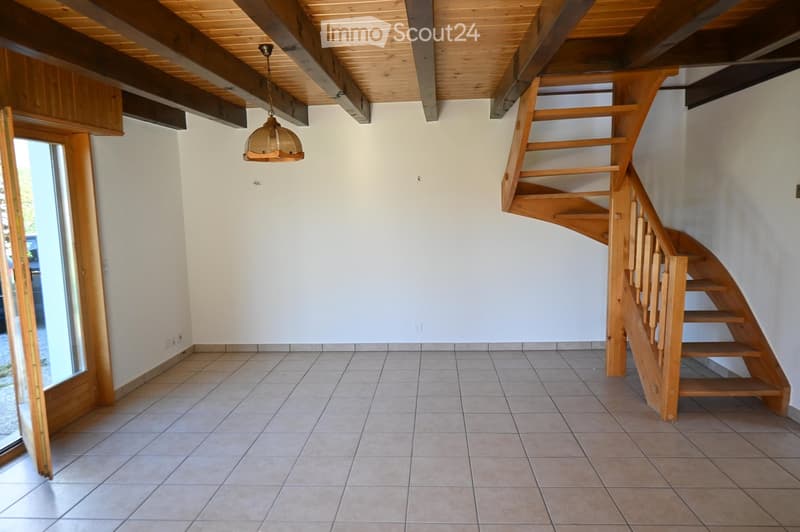 Empty room with wooden staircase, exposed wooden beams on the ceiling, tiled floor, and a hanging light fixture.