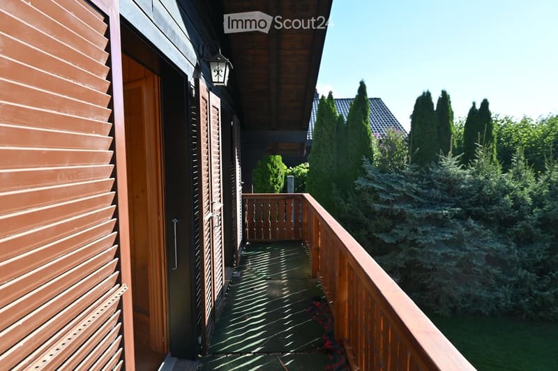 Wooden balcony with railing, adjacent to a building with brown shutters and a wooden door, surrounded by tall evergreen trees.