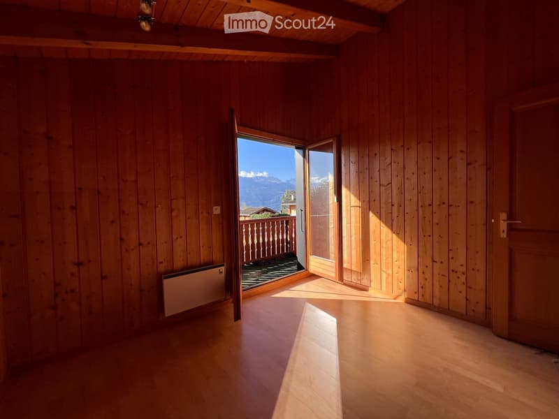 Empty room with wooden walls and floor, large window opening to a balcony with a scenic mountain view.