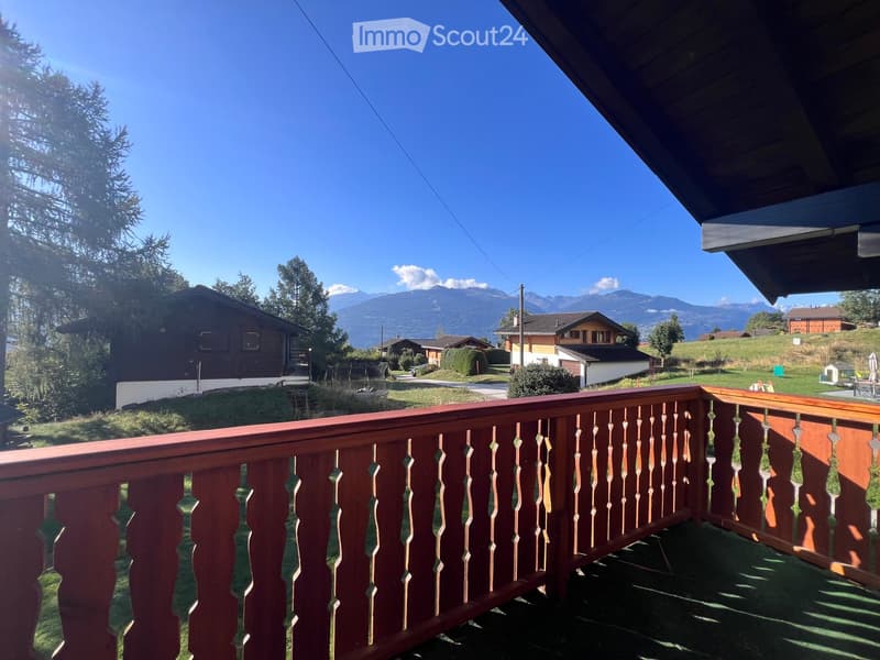 Balcony with wooden railing overlooking a scenic mountain view, surrounded by trees and houses.