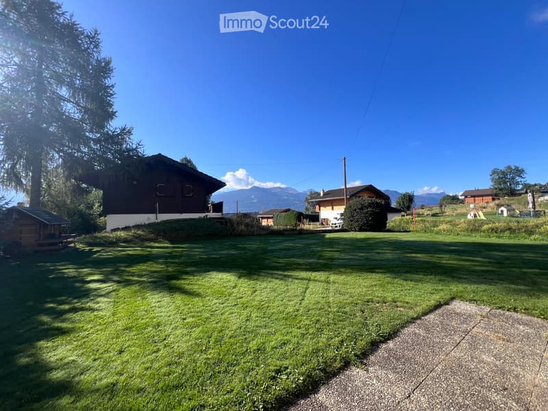 A well-maintained lawn with several houses and trees in the background, under a clear blue sky with distant mountains visible.