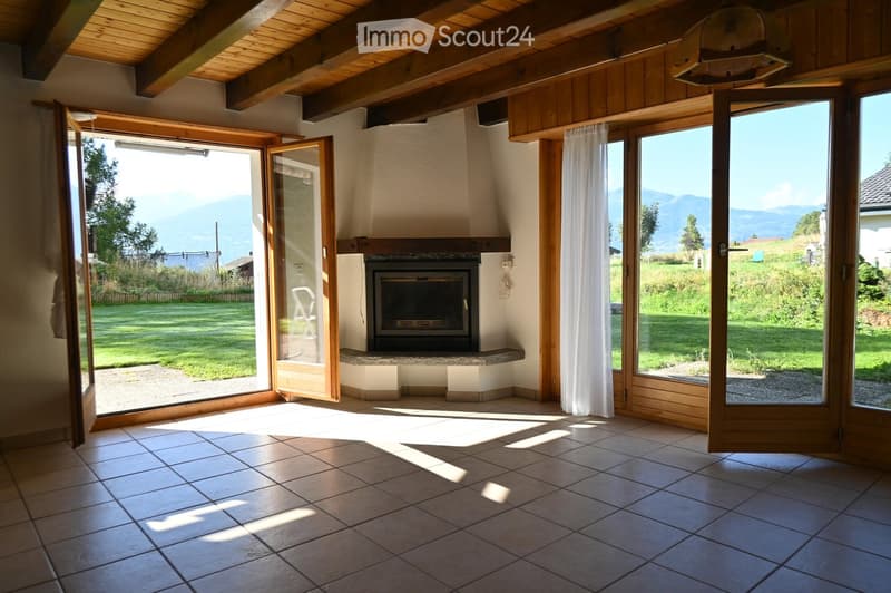 Bright living room with large windows and sliding doors opening to a garden, featuring a central fireplace and wooden beams on the ceiling.