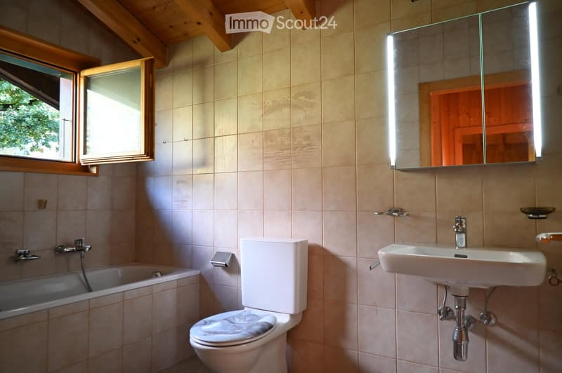 Bathroom with beige tiled walls, a bathtub, toilet, sink, and a large mirror with built-in lighting. The room features wooden beams on the ceiling and a window with wooden frames.