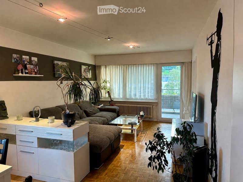 Modern living room with a grey sectional sofa, glass coffee table, white side table, and decorative plants. The room features wooden flooring, large windows with curtains, and a balcony door leading to an outdoor area.