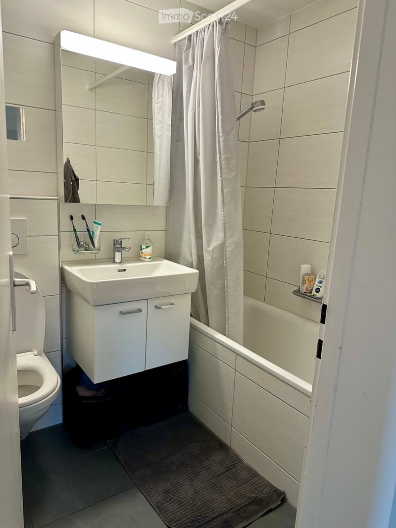 Modern bathroom with white tiles, a sink with a mirror, a toilet, a bathtub with a shower curtain, and a small bathroom cabinet.