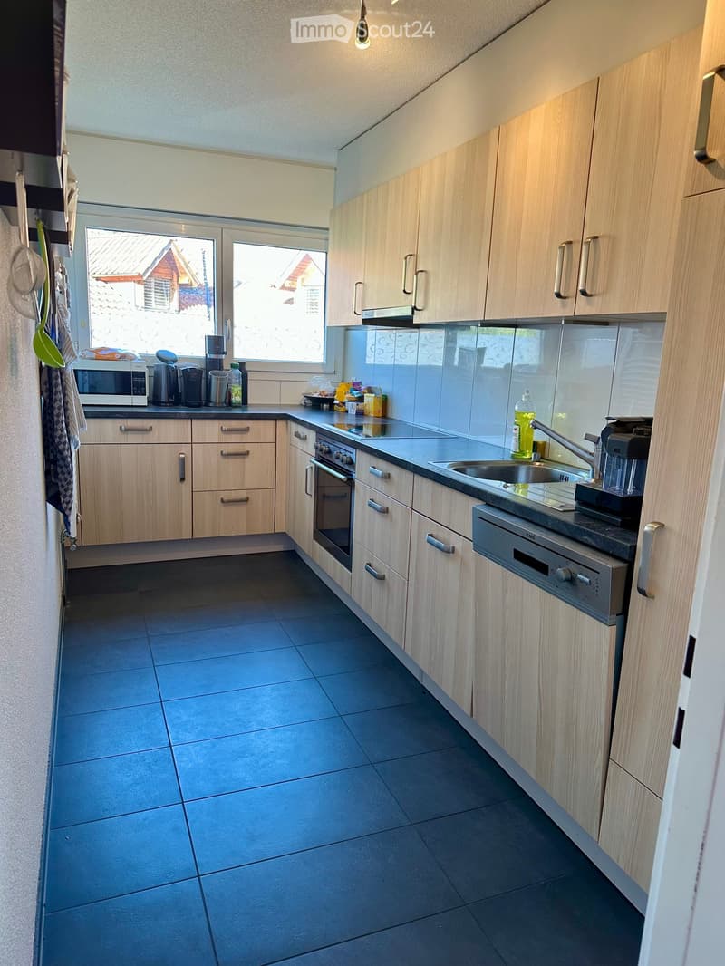 Modern kitchen with light wooden cabinets, stainless steel appliances, and a dark tiled floor.